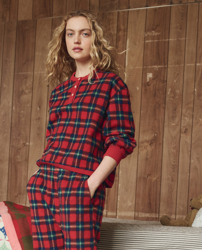A person with long curly hair stands indoors before a wooden wall, wearing THE GREAT. H25 SLEEP's Plush Fleece Henley Sleep Sweatshirt in Cedarwoods Plaid.