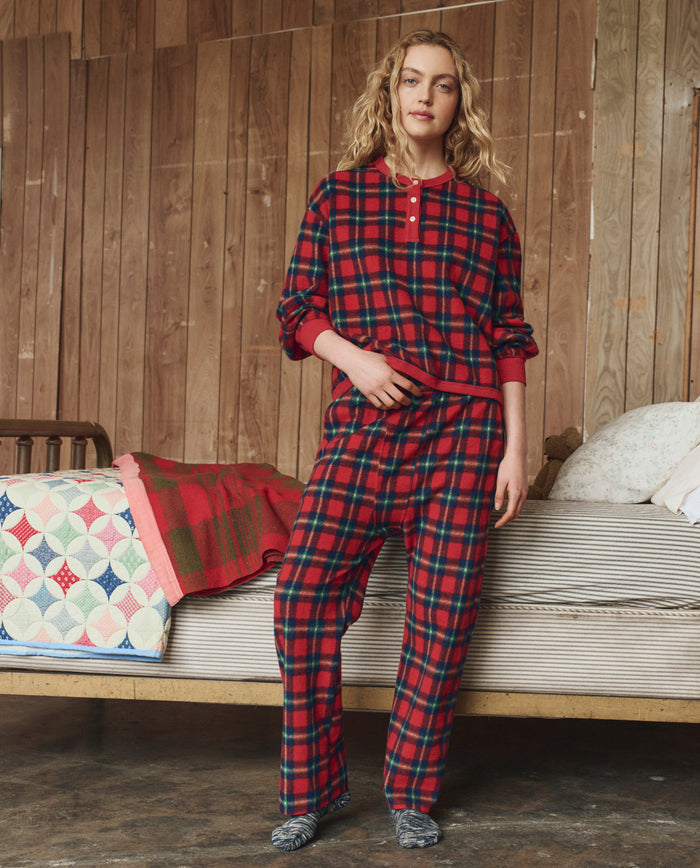 A person wears THE GREAT. H25 SLEEP's The Plush Fleece Henley Sleep Sweatshirt in Cedarwoods Plaid, standing indoors by a quilted bed and wood-paneled wall.