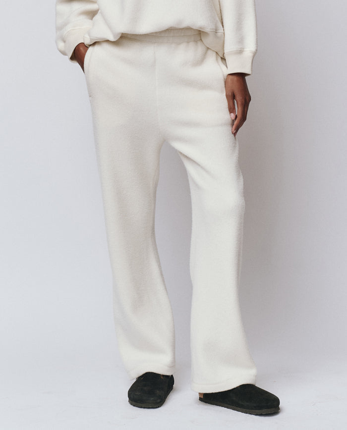 Person wears THE GREAT. H25 TEDDY FLEECE The Plush Victory Sweatpant in Washed White, handcrafted in L.A. from ultra-soft fleece, with hands in pockets against a plain background and black slippers.