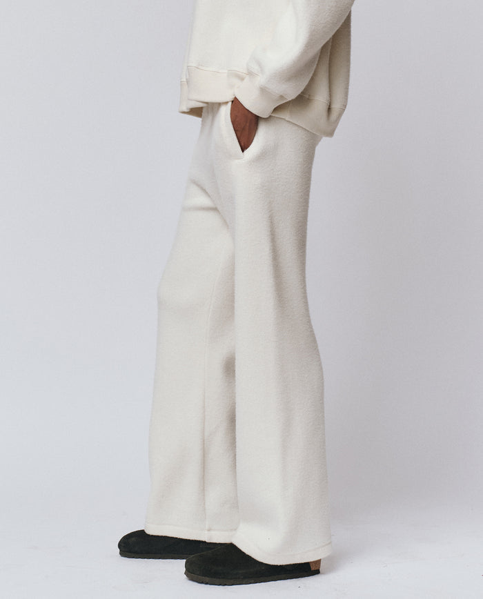 A person wears THE GREAT. H25 TEDDY FLEECE The Plush Victory Sweatpant in Washed White with a matching sweatshirt, one hand in pocket, black shoes visible, standing against a plain background. Handcrafted in L.A.