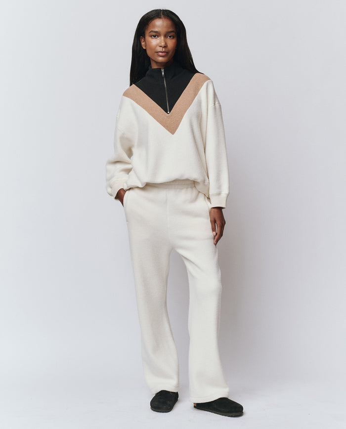 A woman stands against a plain background wearing THE GREAT. H25 TEDDY FLEECE The Plush Victory Sweatpant in Washed White, paired with a matching handcrafted top featuring a beige and black V-shaped design, and black slip-on shoes.
