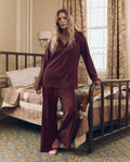 A woman wearing THE GREAT. F26 SLEEP’s “The Wide Leg Sleep Pant” in maroon stands by a bed in a vintage-style bedroom with patterned curtains and a patchwork quilt.
