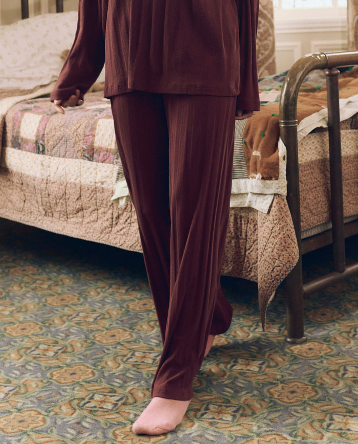 A person wearing THE GREAT. F26 SLEEP The Wide Leg Sleep Pant in maroon and pink socks stands next to a bed with a patchwork quilt in a carpeted room.
