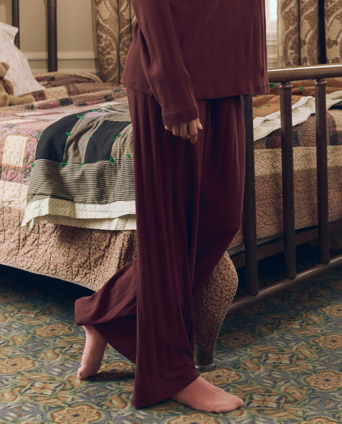 A person stands barefoot beside a bed with a patchwork quilt in a carpeted bedroom, wearing THE GREAT. F26 SLEEP’s The Wide Leg Sleep Pant in Maroon.