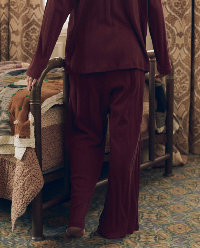 A person wearing THE GREAT. F26 SLEEP The Wide Leg Sleep Pant in maroon stands next to a bed with a patchwork quilt in a patterned, carpeted room with curtains.