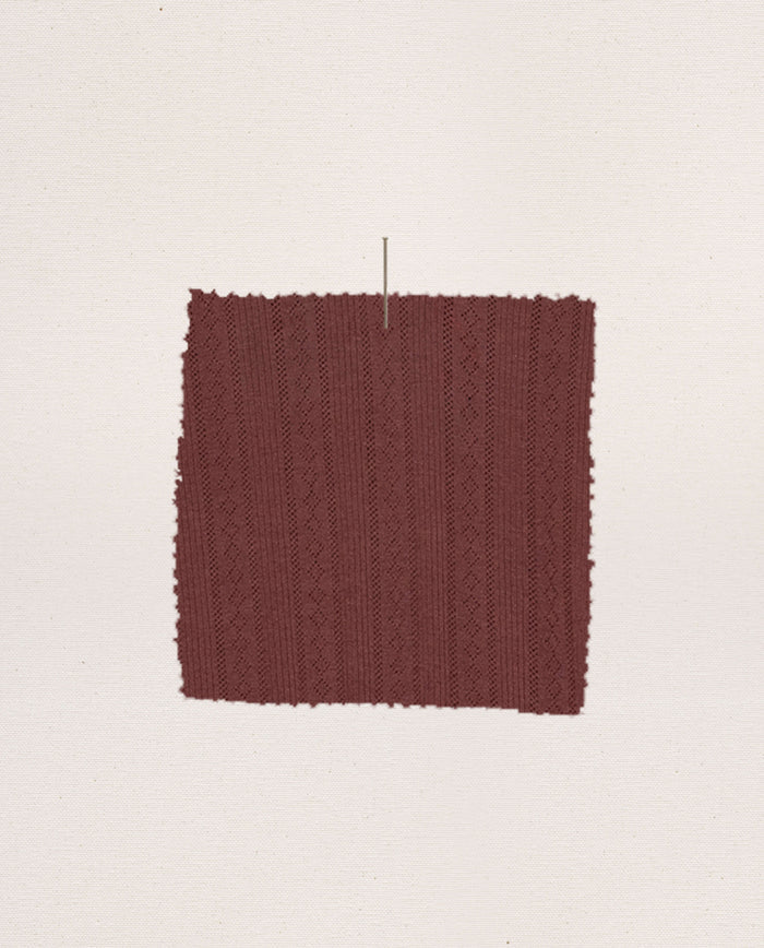 A rectangular swatch of textured maroon fabric with a subtle geometric pattern, from THE GREAT. F26 SLEEP’s The Wide Leg Sleep Pant, is pinned against a plain off-white background—ideal for crafting cozy lounge or wide leg sleep pants.
