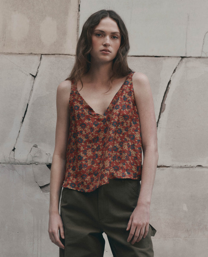 A person with long hair wearing THE GREAT. F26 D1 The Port Cami. -- Gilded Poppy, a red floral sleeveless top, and green pants stands in front of a cracked, beige wall.