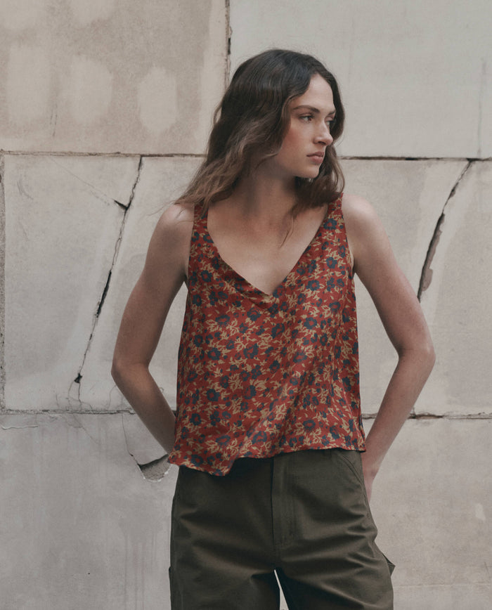 A woman stands against a cracked, light-colored wall, looking to the side, wearing THE GREAT. F26 D1 The Port Cami. in Gilded Poppy and olive green pants.