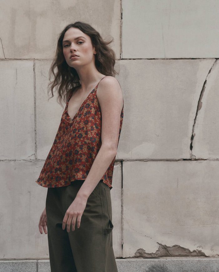 A woman with wavy hair wears The Port Cami in Gilded Poppy by THE GREAT. F26 D1 and olive trousers while standing in front of a weathered stone wall.