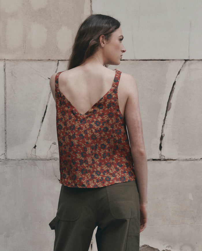 A person stands with their back to the camera, wearing THE GREAT.’s The Port Cami. in Gilded Poppy—a red floral sleeveless top with a deep V back—paired with olive green pants, in front of a textured stone wall.