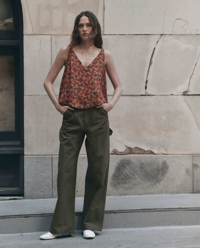 A model stands outdoors, hands in pockets, against a stone wall, wearing THE GREAT. The Port Cami in Gilded Poppy—a red floral top—paired with olive wide-leg trousers and white flats.
