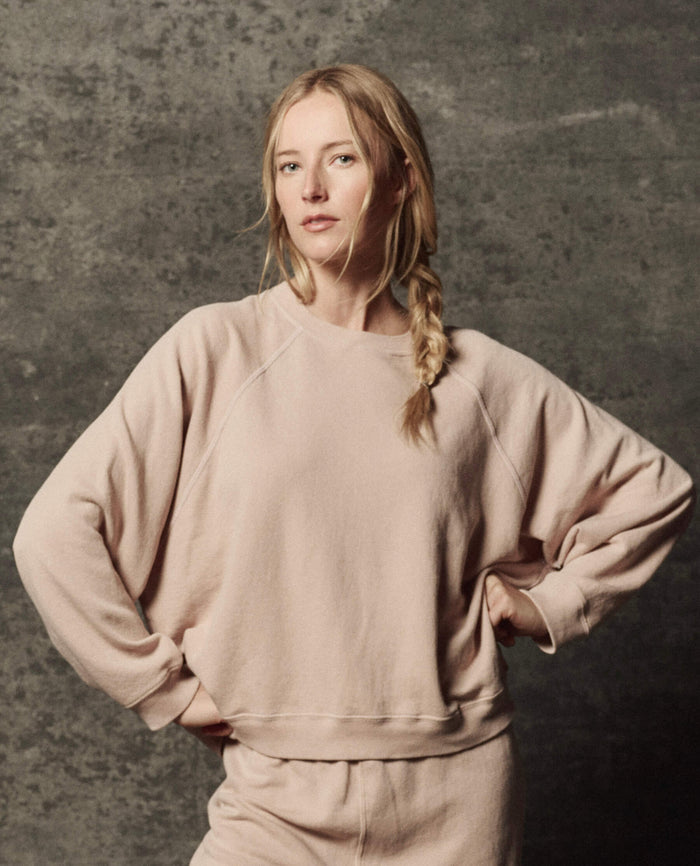 A woman with blonde hair in a side braid stands with hands on hips against a textured gray backdrop, wearing THE GREAT. PF26 DANCER KNIT BODIES' The Rehearsal Sweatshirt in Ballet Pink, paired with matching pants.