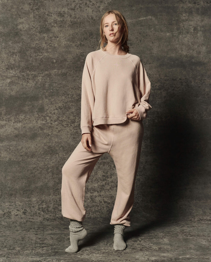 A person stands on one leg, wearing THE GREAT. PF26 DANCER KNIT BODIES' The Rehearsal Sweatshirt in Ballet Pink with matching pants and gray socks, against a gray textured background.