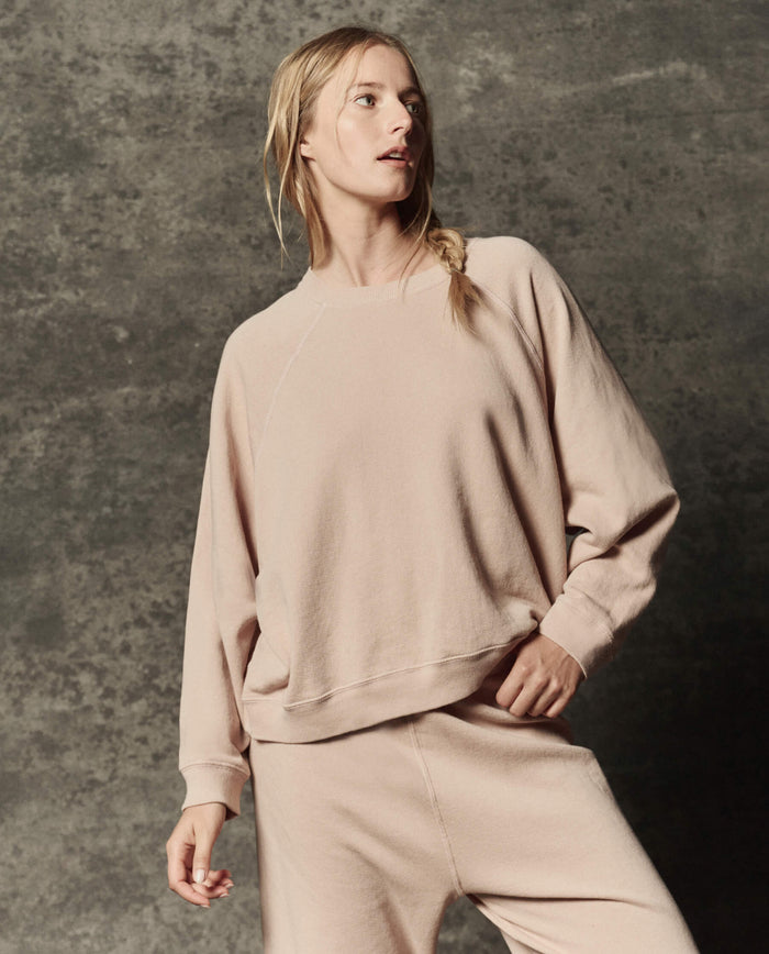 A person dressed in THE GREAT. PF26 DANCER KNIT BODIES The Rehearsal Sweatshirt in Ballet Pink and matching pants stands against a textured, gray backdrop, looking to the side.