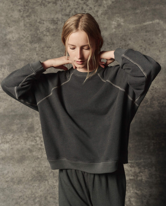 A woman stands against a textured gray background in THE GREAT. PF26 DANCER KNIT BODIES' The Rehearsal Sweatshirt in Flat Charcoal, looking down as she lifts her hands to adjust the collar.