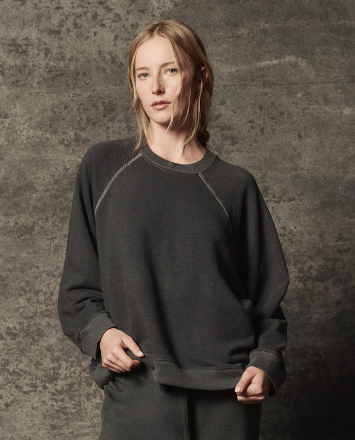 A woman wearing THE GREAT. PF26 DANCER KNIT BODIES' The Rehearsal Sweatshirt in Flat Charcoal and matching pants stands in front of a textured gray background, gazing at the camera with a neutral expression.