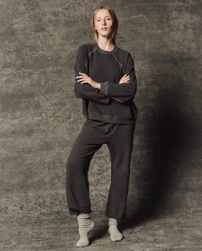Standing against a textured gray background, the person wears THE GREAT. PF26 DANCER KNIT BODIES' The Dance Pant in Flat Charcoal, paired with a loose dark sweatshirt and light gray socks. Their arms are crossed, and they hold a neutral expression.
