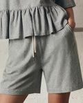 A person wearing THE GREAT. SP26 DANCER BODIES Relaxed Sport Short in Heather Grey stands against a light textured wall with one hand in their pocket, paired with a grey ruffled top.