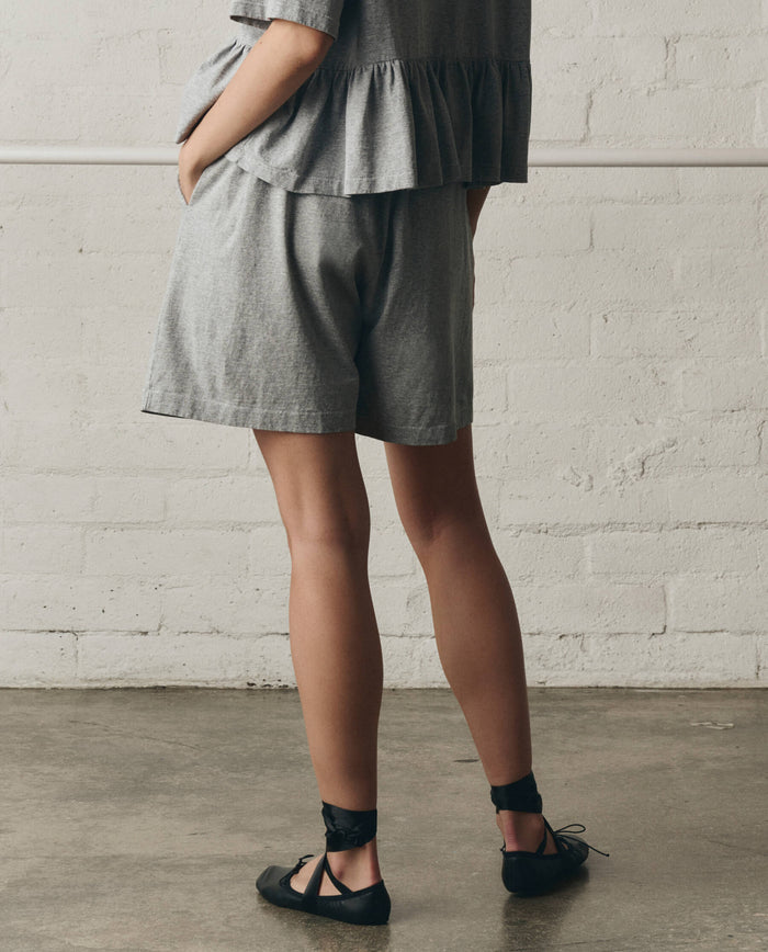A person stands indoors against a white brick wall, wearing THE GREAT. SP26 DANCER BODIES’ The Relaxed Sport Short in Heather Grey with a grey heavyweight jersey short-sleeve top and black ballet flats with ankle ties.