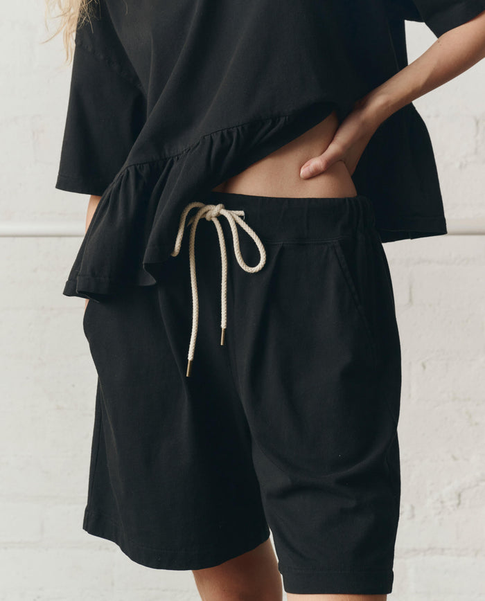Model wears THE GREAT.'s Relaxed Sport Short in True Black, paired with a loose black short-sleeve top; hands in pockets highlight the relaxed fit against a light background.