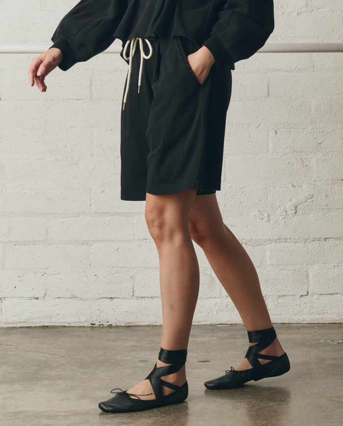 Someone wearing The Relaxed Sport Short in True Black by THE GREAT. SP26 DANCER BODIES, paired with a black heavyweight jersey long-sleeve top with a drawstring waist and black lace-up ballet flats, stands indoors against a white brick wall.