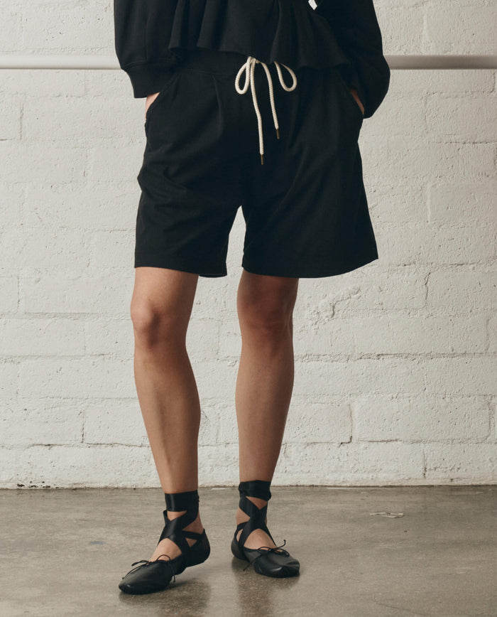 Wearing THE GREAT. SP26 DANCER BODIES The Relaxed Sport Short in True Black, paired with a black heavyweight jersey long-sleeve top and black ballet flats with ribbon ankle ties, standing indoors against a white brick wall.