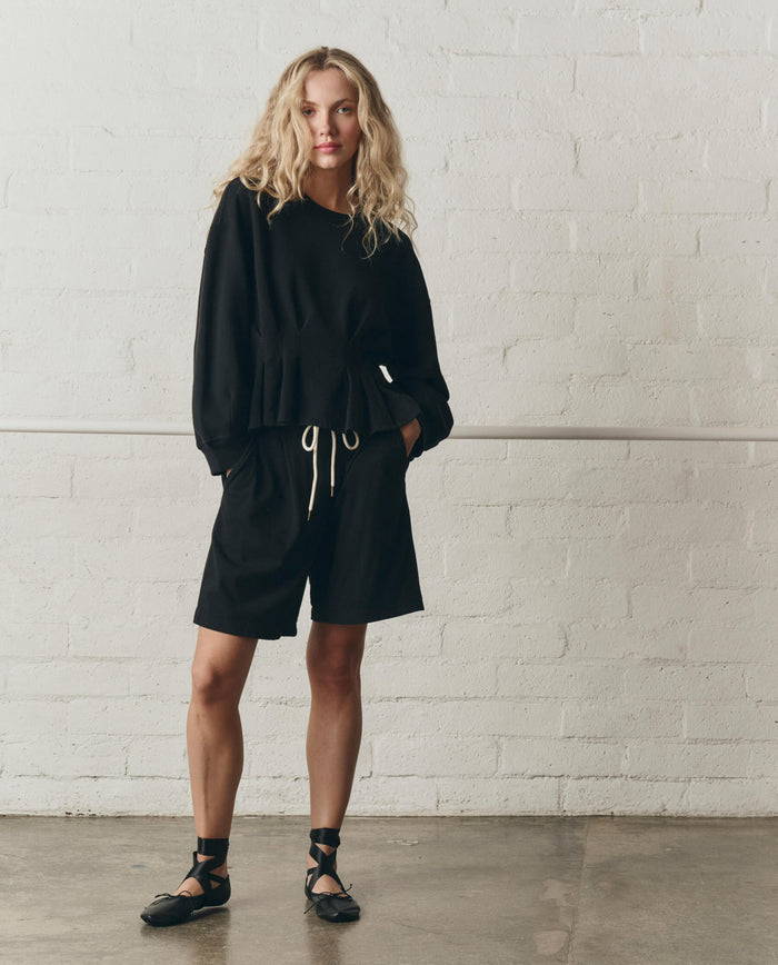 A person stands indoors against a white brick wall, wearing THE GREAT.'s SP26 DANCER BODIES black long-sleeve top and The Relaxed Sport Short in True Black, paired with black ballet-style shoes.