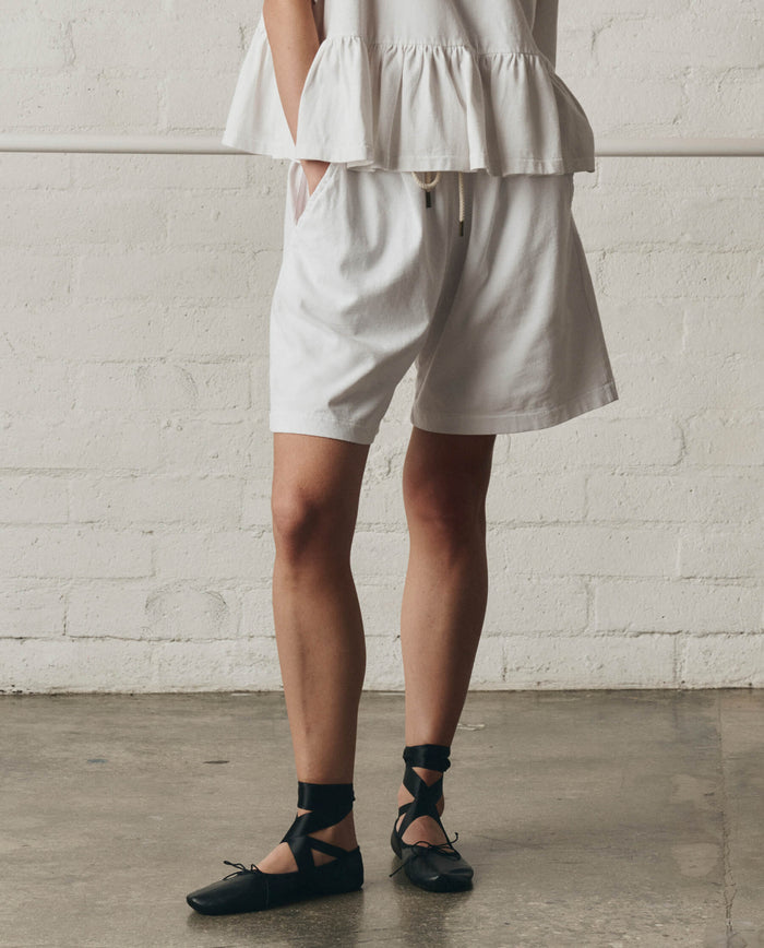 Indoors, a person wears THE GREAT. SP26 DANCER BODIES’ The Relaxed Sport Short in True White—featuring an elastic waistband and pockets—with a white ruffled top and black lace-up flats. The backdrop is a light brick wall and concrete floor.