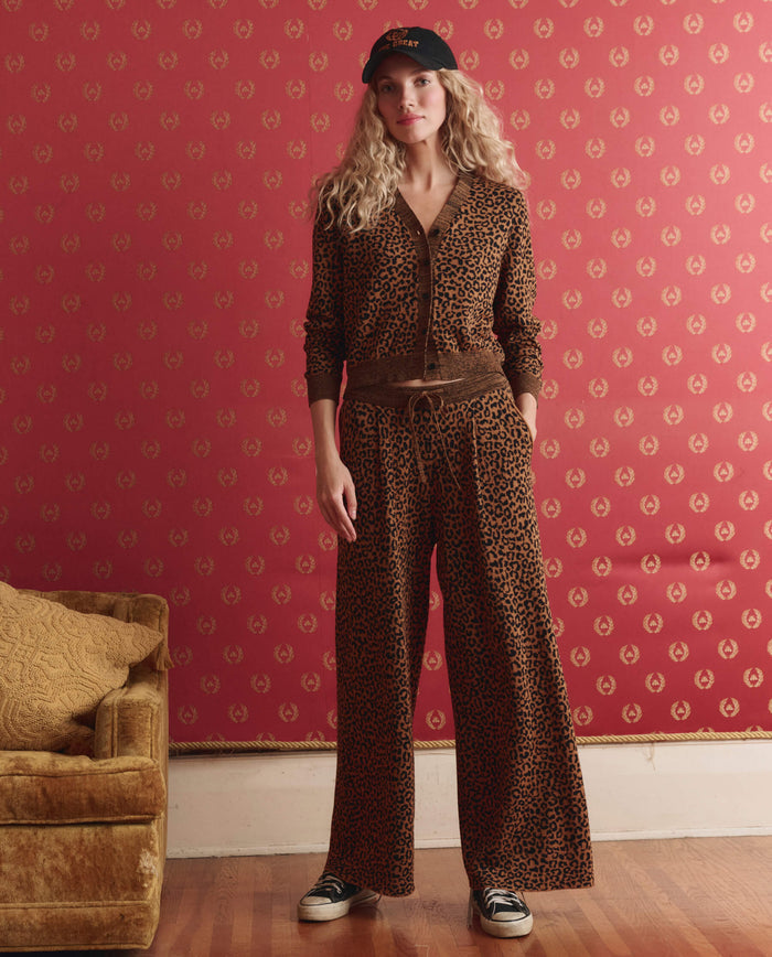 A woman stands indoors against a red patterned wall, wearing THE GREAT. SP26 D1 The Relaxed Trouser Pant in Leopard, paired with a black cap and sneakers, with one hand in her pocket.