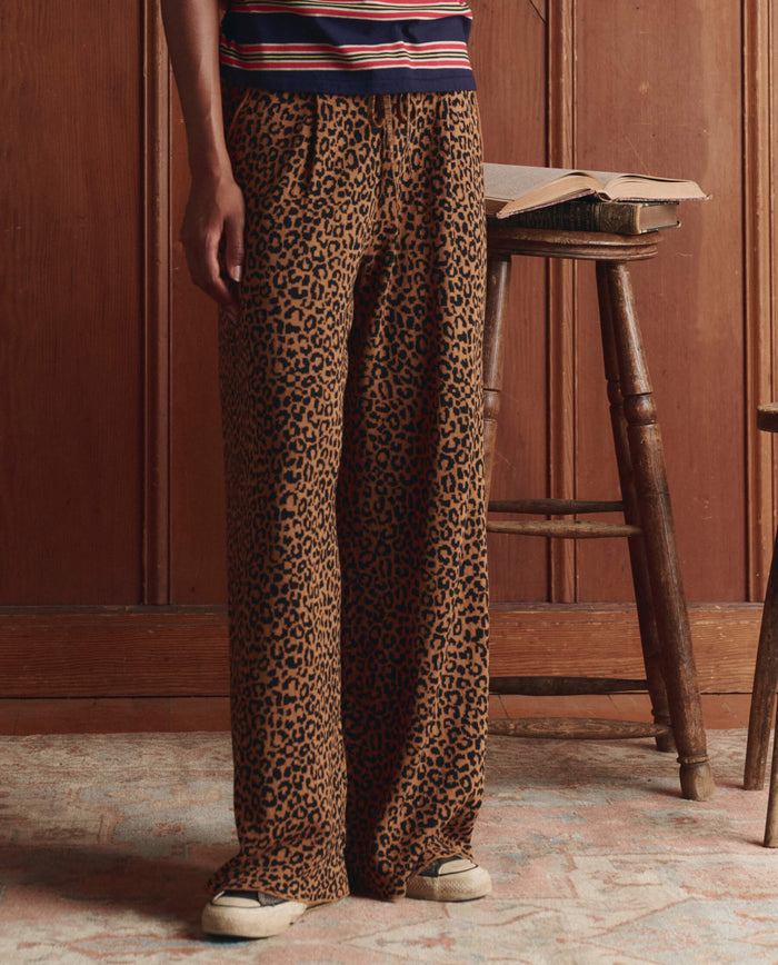 A person indoors wears THE GREAT. SP26 D1 The Relaxed Trouser Pant in leopard print with sneakers, resting a hand on a wooden stool topped with an open book.