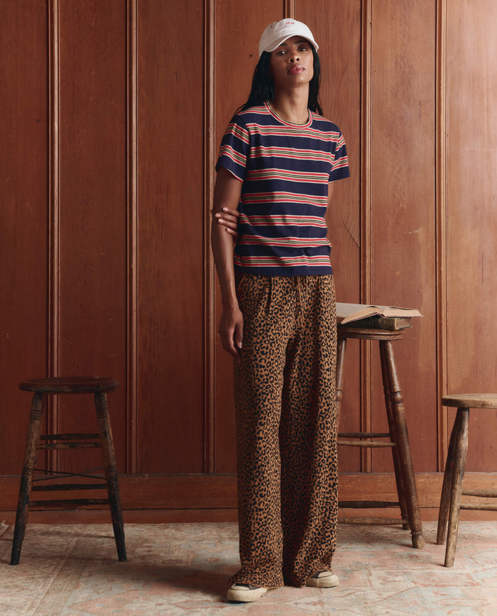 A person wearing The Little Tee in Scout Stripe by THE GREAT. SP26 D1 stands indoors between two wooden stools against a wood-paneled wall.