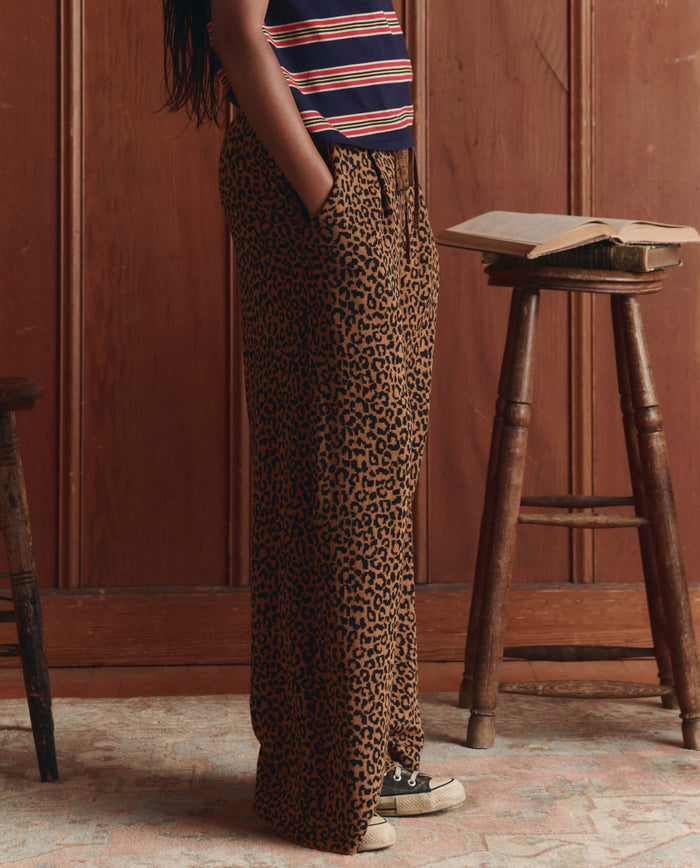 A person in THE GREAT. SP26 D1 The Relaxed Trouser Pant—Leopard, black sneakers, and a striped top stands by a wooden stool with an open book in a wood-paneled room.