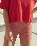 Wearing THE GREAT. SU26 Relay Short in Pimento Stripe, the person stands outdoors with parts of their arm and legs visible.