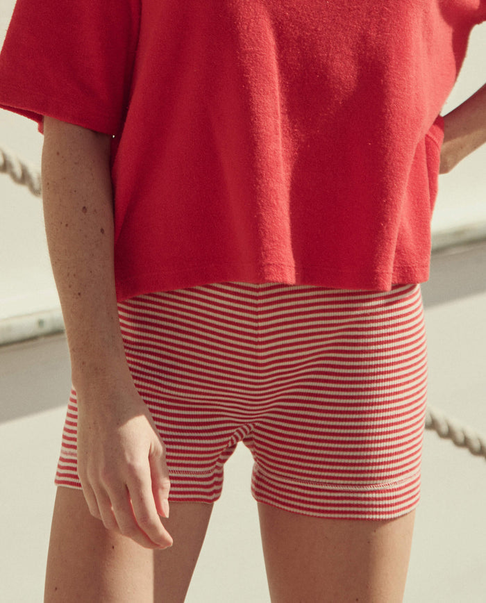Wearing THE GREAT. SU26 Relay Short in Pimento Stripe, the person stands outdoors with parts of their arm and legs visible.