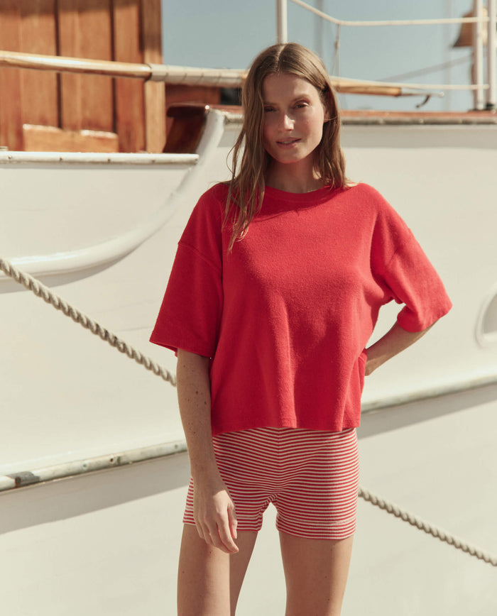 A woman stands outdoors in front of a white boat, wearing a red short-sleeve top and THE GREAT. SU26's The Relay Short in Pimento Stripe.