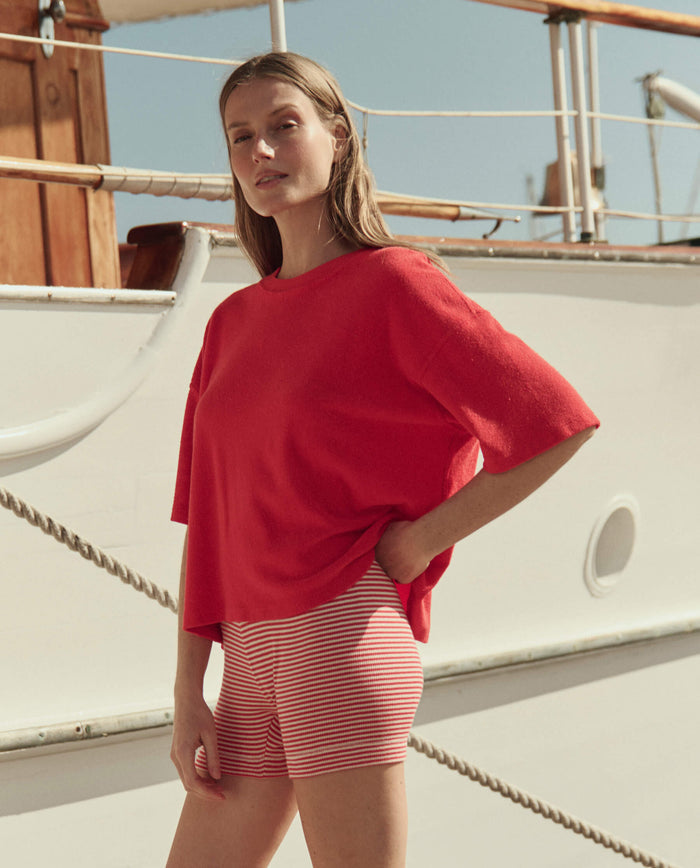 A woman wearing THE GREAT. SU26’s The Relay Short in Pimento Stripe stands in front of a white boat on a sunny day, paired with a loose red top.