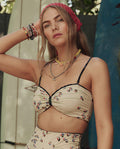 A woman with long hair in a red bandana wears THE GREAT. SP26 SWIM’s Windblown Bouquet Reversible Cinched Bandeau, layered with necklaces, outdoors with a surfboard in the background.