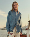 A woman stands outdoors near a marina, wearing THE GREAT. SU26 The Road Shirt -- Shoreline Wash and light-colored pants with red and green floral embroidery.