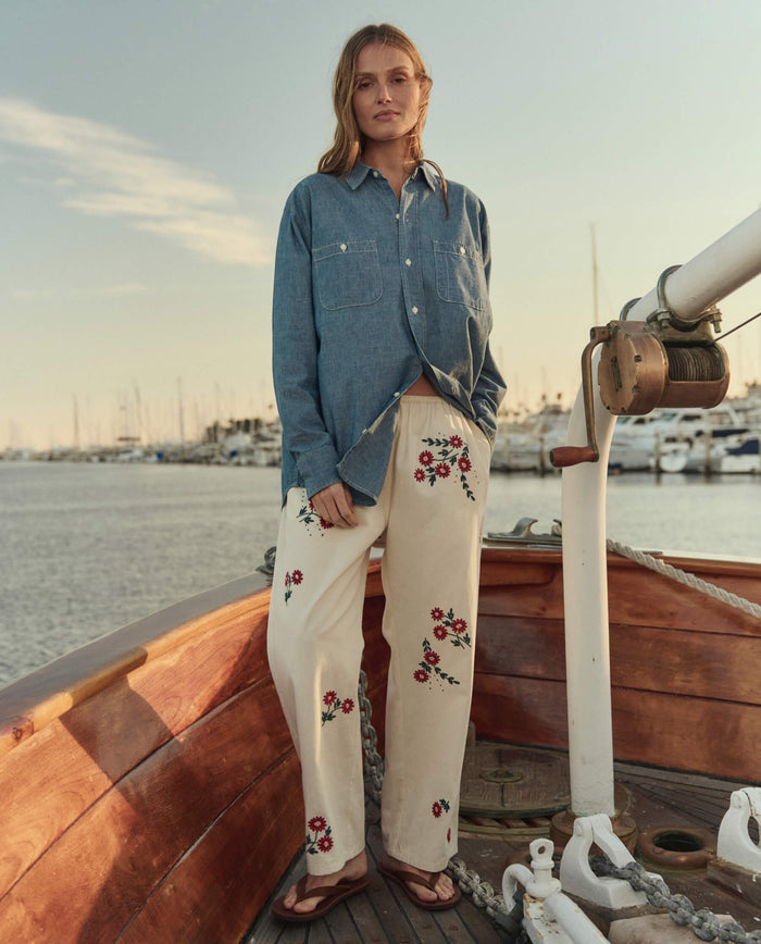 At a marina, someone stands on a wooden boat wearing THE GREAT. SU26 The Road Shirt in Shoreline Wash, cream pants with red floral embroidery, and sandals.
