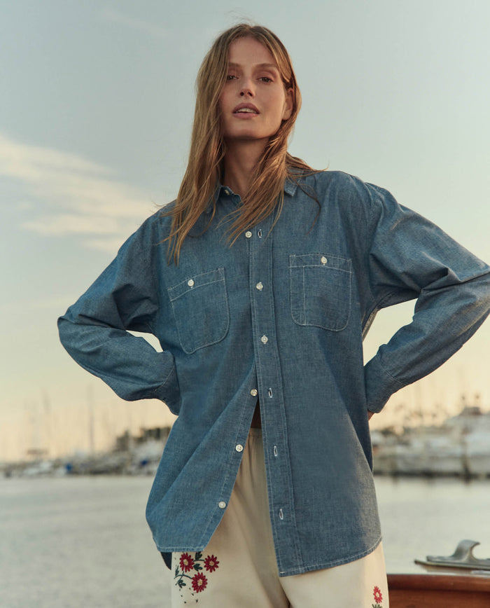 A woman stands outdoors near a marina, wearing THE GREAT. SU26 The Road Shirt in Shoreline Wash and cream-colored pants adorned with red floral embroidery.