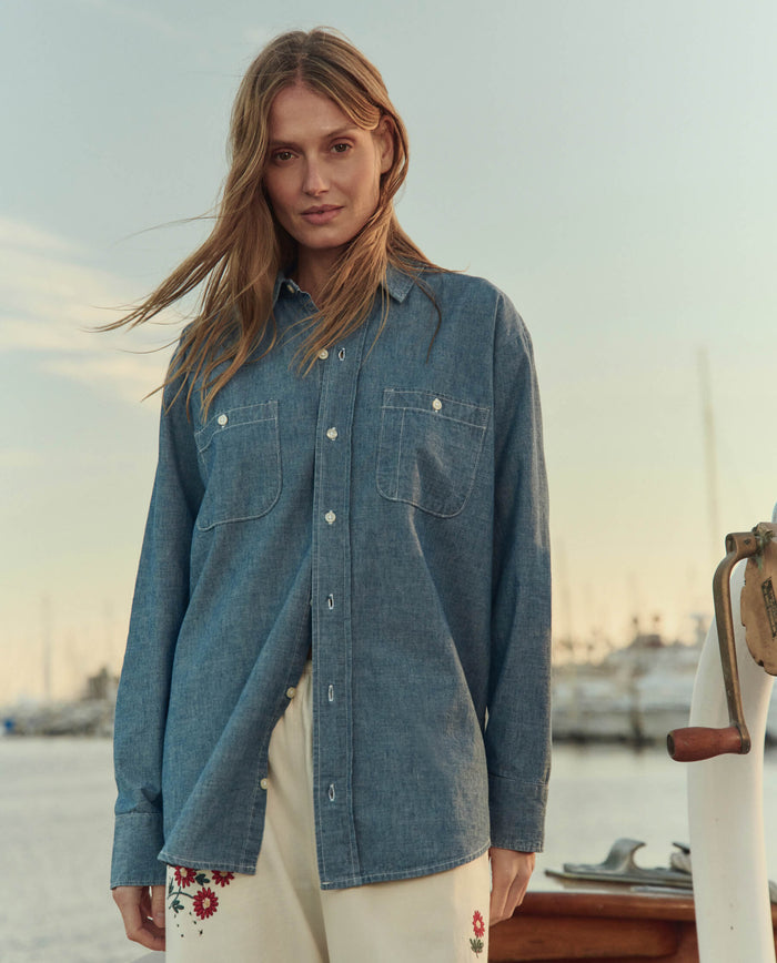 A woman stands outdoors near a marina wearing THE GREAT. SU26 The Road Shirt in Shoreline Wash and white pants with red floral embroidery.