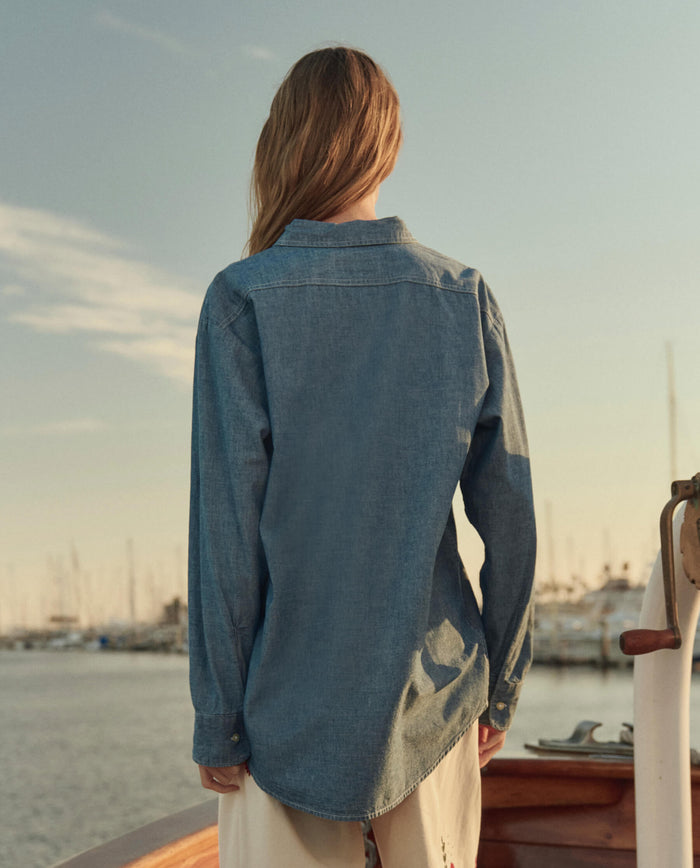 A person with long hair, seen from behind, wears THE GREAT. SU26 The Road Shirt - Shoreline Wash and light pants while standing on a boat with a marina and sailboats in the background.