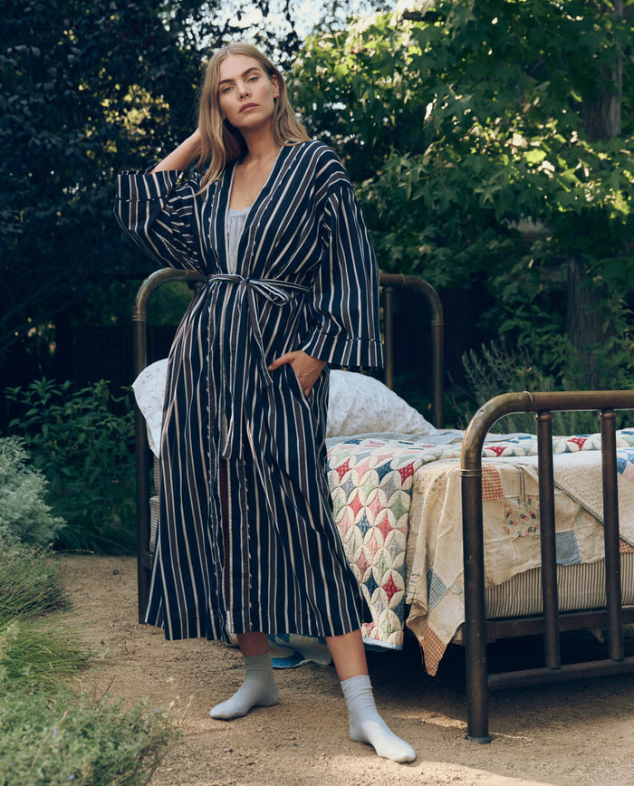 A woman in The Great. SP26 Sleep's The Robe. -- Coastline Stripe stands barefoot by an outdoor metal bed with a patchwork quilt, surrounded by lush greenery.