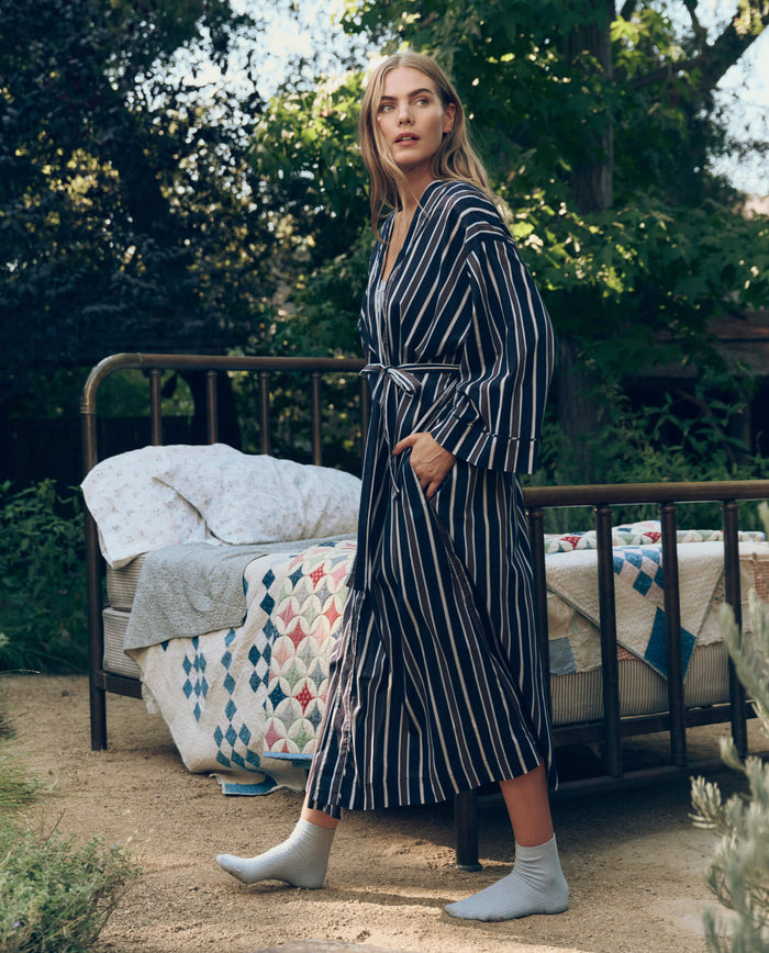 A woman in The Great. SP26 Sleep’s The Robe. -- Coastline Stripe stands outdoors by a metal bed with patchwork quilts, surrounded by greenery.
