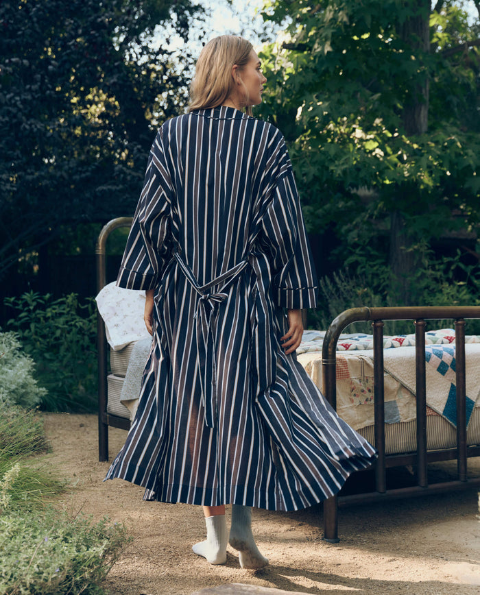 Wearing The Robe in Coastline Stripe by The Great. SP26 Sleep, a person stands outdoors beside a metal bed frame with quilts, surrounded by greenery.