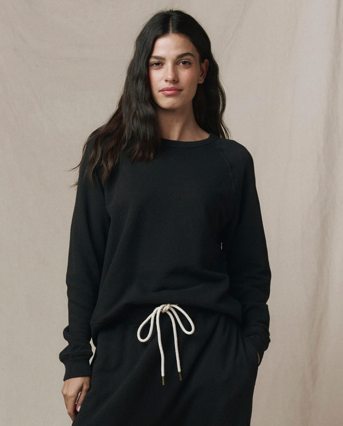 A person with long dark hair wears the SP26 New Knits "The Rookie Sweatshirt. Solid -- Almost Black" and matching black pants with a white drawstring, standing in front of a plain beige backdrop.