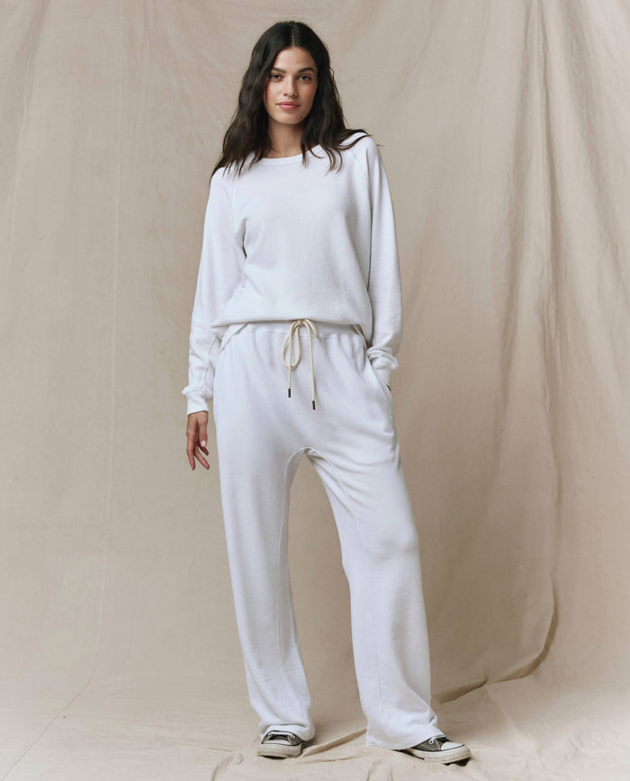 A woman stands against a beige backdrop, wearing the SP26 New Knits The Rookie Sweatshirt in True White with matching drawstring pants and white sneakers.