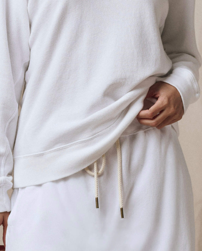 A person wears SP26 New Knits' The Rookie Sweatshirt in Solid -- True White, paired with matching white drawstring pants, holding the sweatshirt’s hem with one hand.
