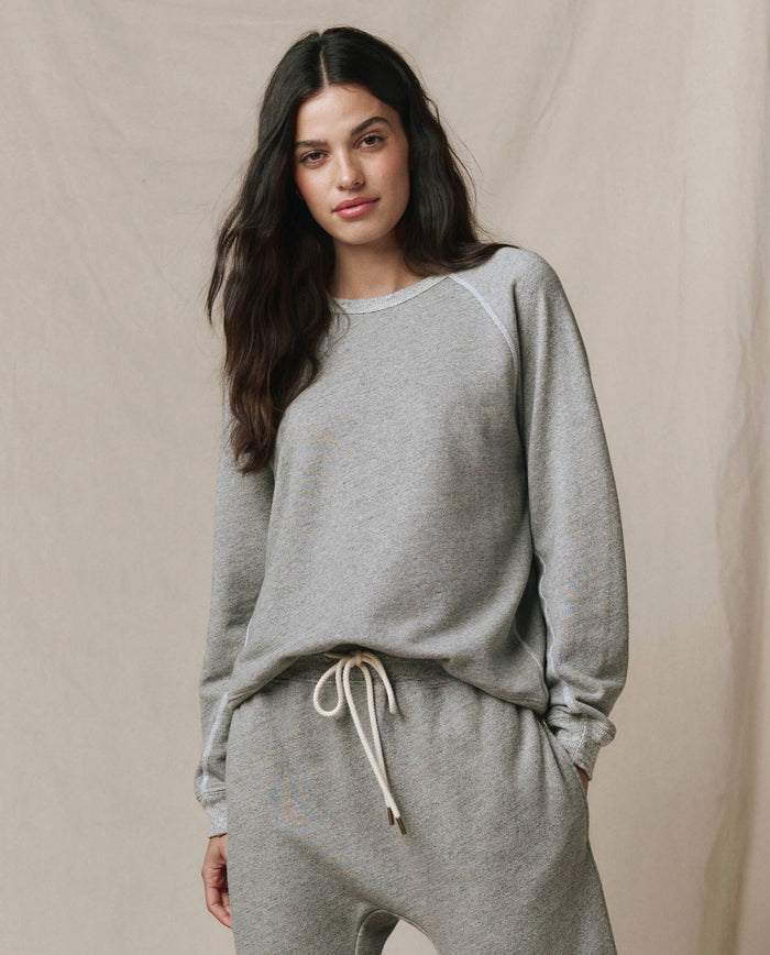 A woman models the SP26 New Knits "The Rookie Sweatshirt" in Varsity Grey, paired with matching drawstring sweatpants, standing before a neutral fabric backdrop.