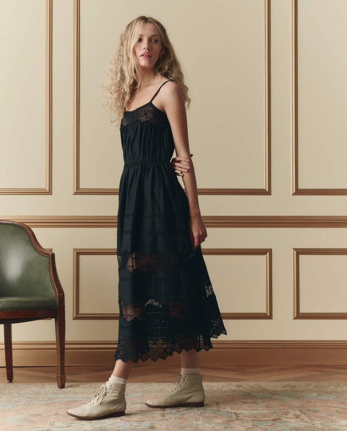 A woman with long curly hair wears THE GREAT. SP26 D2’s The Rosette Crochet Dress in black and cream lace-up boots, standing beside a green chair in a room with paneled walls.